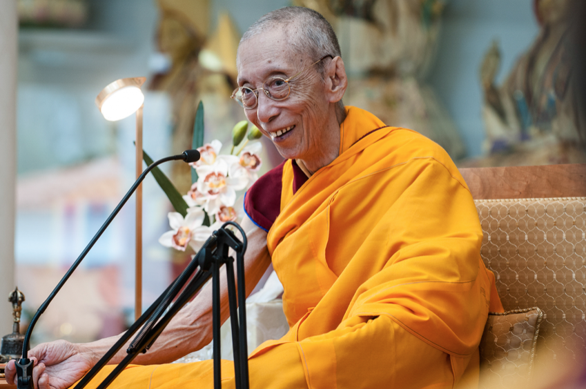 Moine bouddhiste souriant vêtu d’une robe orange, assis avec un micro et des fleurs à l’arrière-plan.